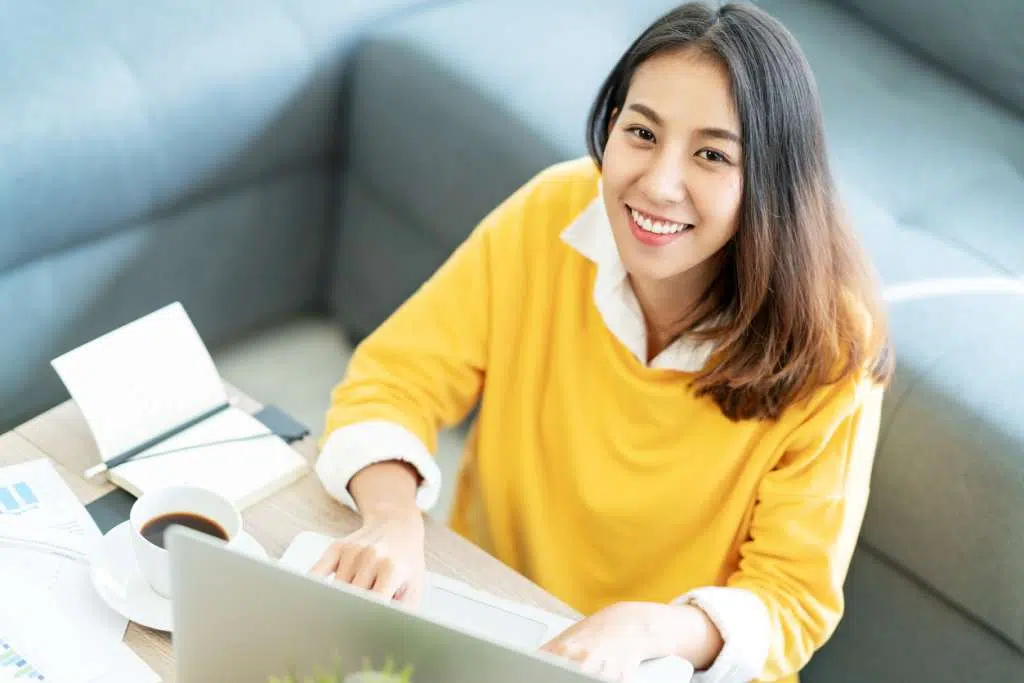 Young attractive happy asian female student sitting at living room floor smiling and looking up at camera working on laptop at home office. Young startup entrepreneur or freelance business concept.